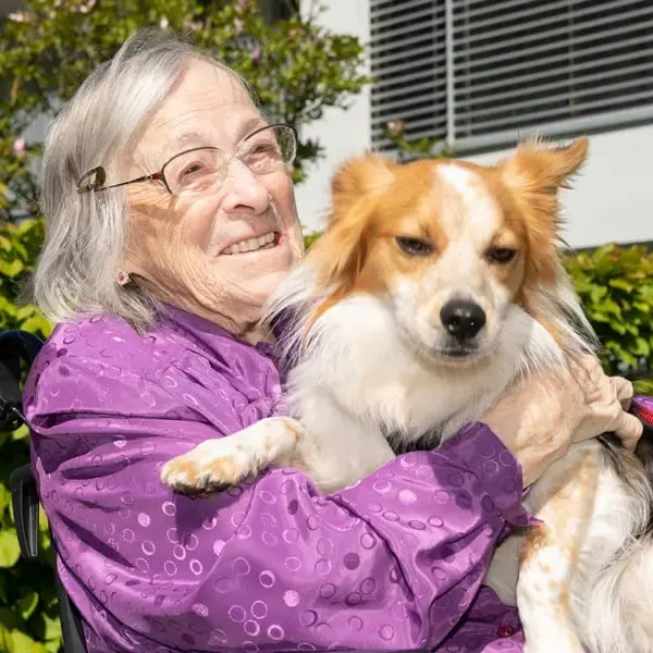 Eine ältere Frau hält draußen ihren weiß-braunen Hund im Arm und lächelt dabei herzlich