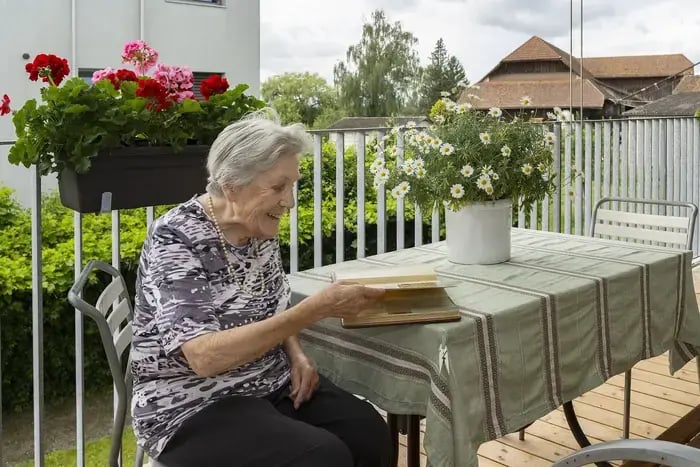 Ältere Frau liest ein Buch an einem Gartentisch, umgeben von Blumen