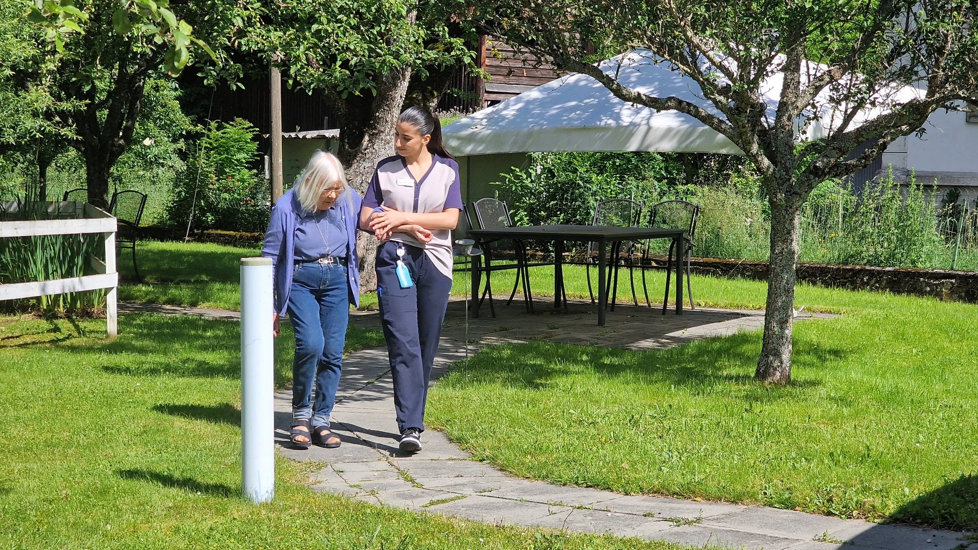 Résidente accompagnée d’une soignante marchant dans le jardin verdoyant de Tertianum La Fontaine