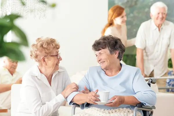 Zwei alte Damen lachen und plaudern und trinken ihren Kaffee, im Hintergrund ist eine Krankenschwester auf dem Arm eines alten Mannes und sie lachen und plaudern